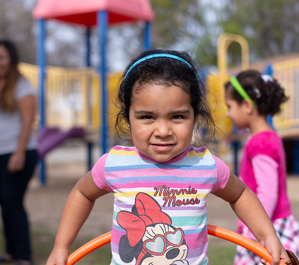 small girl wearing a minnie mouse shirt smiles at the camera while playing with a hula hoop outside