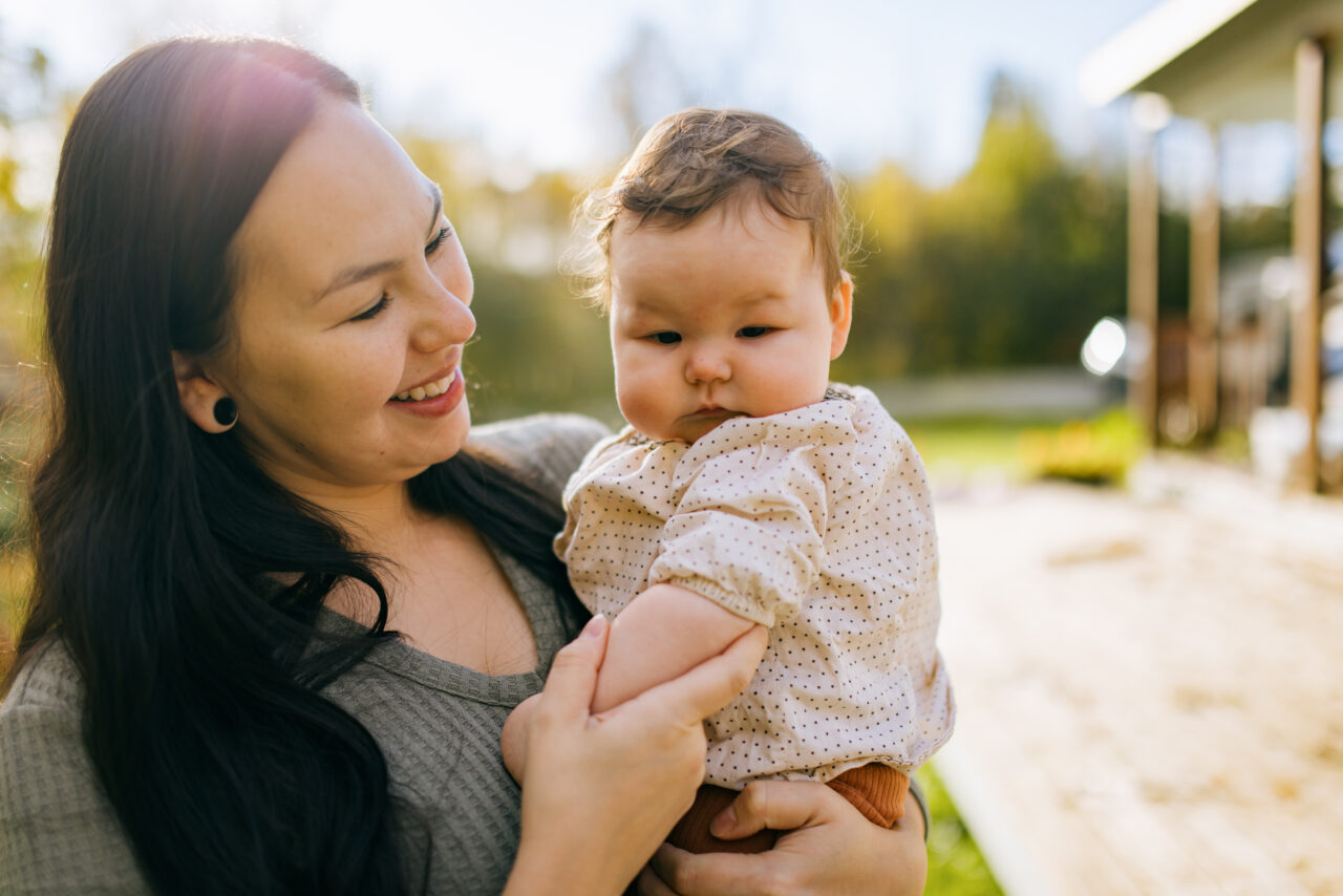 An Indigenous North American woman spends time bonding with her sweet baby girl in their yard, the house visible in the background.